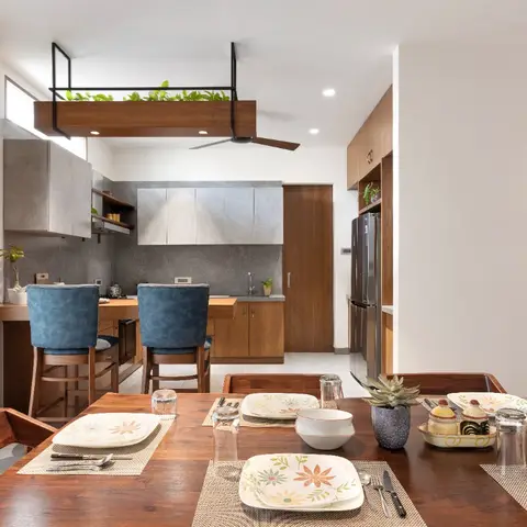 Open dining table set for a meal with a modular kitchen and blue-upholstered breakfast counter stools visible behind it.