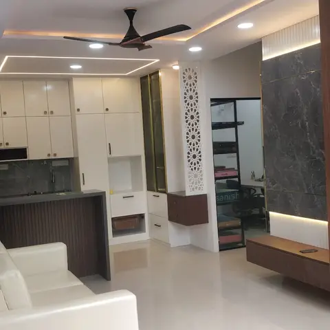 Open kitchen-and-living space with white gloss kitchen cabinets, a carved jaali screen, and a dark marble-and-wood-slat TV wall beside a white sofa.