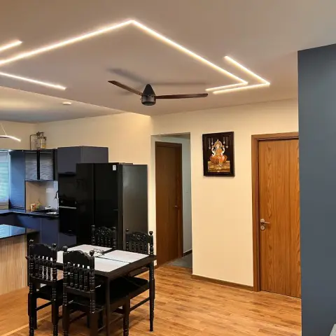 Open kitchen-dining lit by a zig-zag of recessed LED profiles, with a navy modular kitchen and fluted breakfast counter on one side and a dark carved wooden