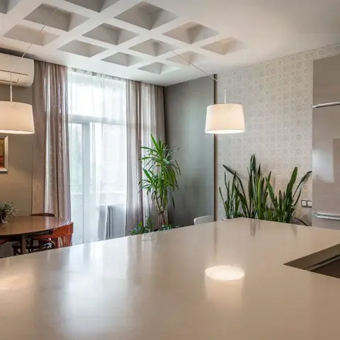 Open kitchen-dining space with a large cream quartz island counter in the foreground, a round wood dining table with rattan-back chairs by a curtained window