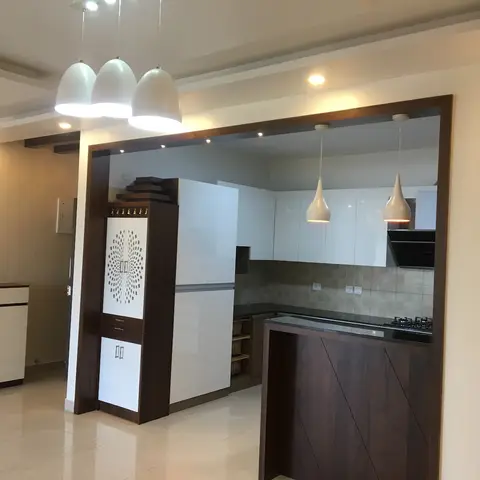 Open kitchen framed by a dark walnut archway with a built-in jaali pooja unit and white gloss cabinetry beyond