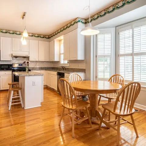Open kitchen with white raised-panel cabinets, stainless appliances, and a granite-topped island beside a round oak breakfast table in a bay-window nook.