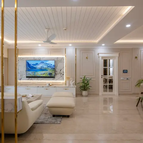 Open living area with a marble-clad TV wall framed by brass rods, fluted wall panelling, a cove-lit stepped ceiling, and cream marble flooring throughout.