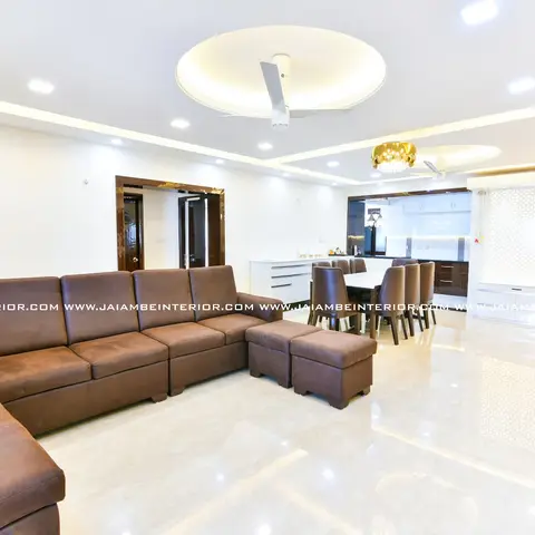 Open-plan apartment shot showing a brown sectional sofa in the foreground, a dining table under a crystal pendant, a kitchen counter