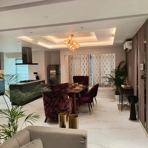 Open-plan apartment showing a dark modular kitchen at the back, a marble-top dining table with maroon floral and velvet chairs under a glass bubble chandelier