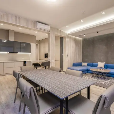 Open-plan apartment showing a grey dining set in the foreground, galley kitchen behind, and a blue corner sofa against a dark stone-effect wall