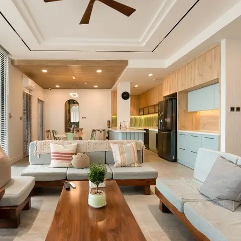 Open-plan apartment showing a grey sectional living area, dining table and a light-blue modular kitchen with a wood-clad ceiling band in one continuous view