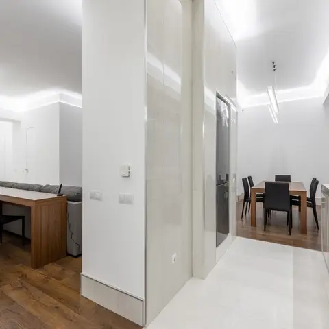 Open-plan apartment showing a living nook with a grey sofa and desk, a dark wood dining table with black chairs, and a white glossy kitchen counter