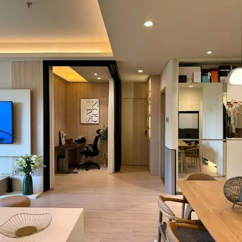 Open-plan apartment view showing a living TV wall, a home-office nook through a black-framed opening, and a dining table with rattan-shaded pendant light