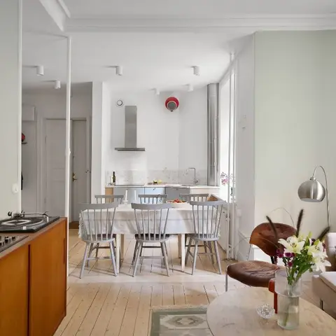Open-plan apartment with a beige sofa and leather sling chair around a round coffee table in the foreground, a dining table with grey spindle chairs behind