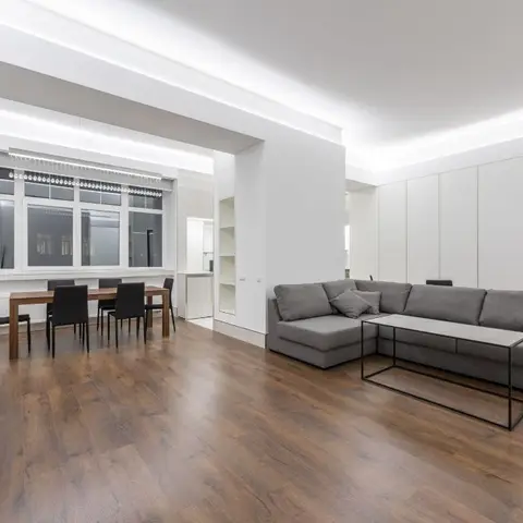 Open-plan apartment with a grey L-shaped sofa and metal-frame coffee table in the foreground and a wood dining table with black chairs under a window in the