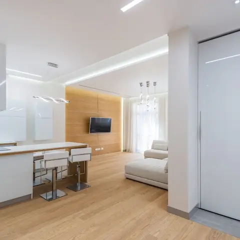 Open-plan apartment with a white kitchen island and bar stools in the foreground and a wood-panelled living area with wall-mounted TV visible beyond