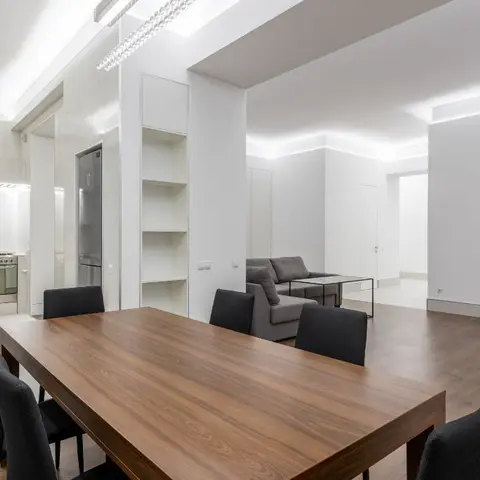 Open-plan apartment with a wood dining table in the foreground, a galley kitchen to the left, and a grey sofa lounge beyond