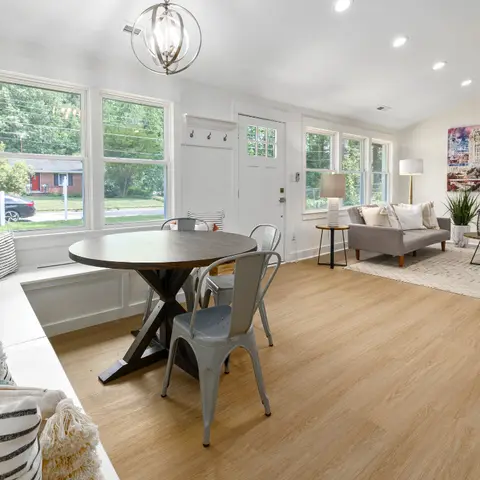 Open-plan dining and living area with a round wood trestle table and metal chairs under an orb chandelier, a built-in window bench with striped pillows