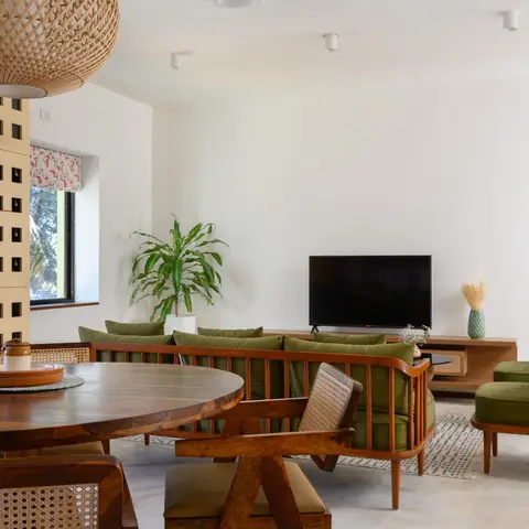 Open-plan dining and living area with a round wood table and cane-back chairs in the foreground, a green bench-style sofa and wood TV console beyond