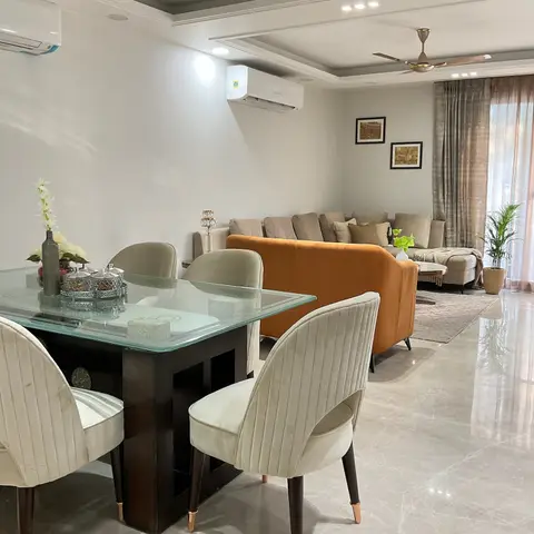 Open-plan dining and living area with a glass-top dining table in the foreground and a burnt-orange velvet sofa against beige curtains beyond.