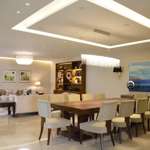 Open-plan dining and living area under a cove-lit ceiling, with a ten-seat dining table beneath a fringe chandelier and a built-in display cabinet dividing it