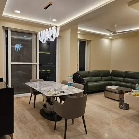 Open-plan dining and living space at night with a sculptural ribbon pendant over a marble-top dining table and a dark green leather sectional beyond.