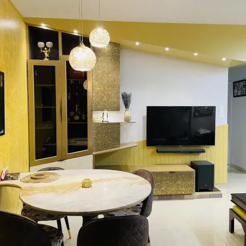 Open-plan dining and living space with a bold mustard wallpaper wall, a round marble dining table with quilted chairs, a brass-textured TV wall unit