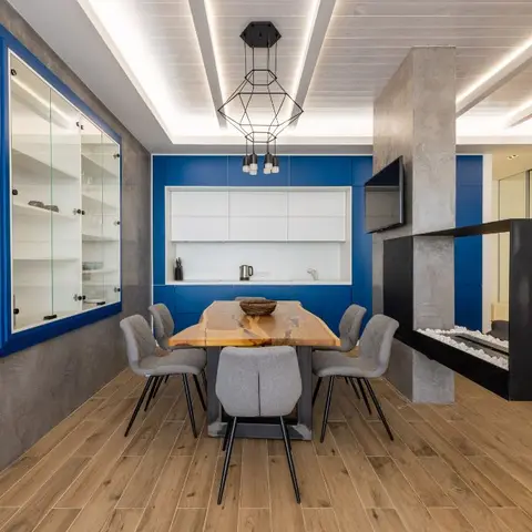 Open-plan dining area with a live-edge wood table and grey chairs in front of a cobalt-blue kitchen wall, textured concrete pillar with built-in fireplace