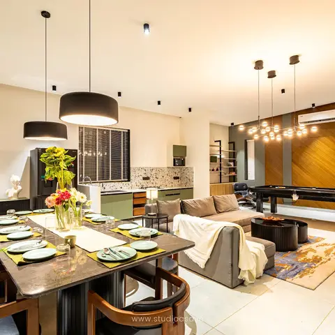 Open-plan dining, kitchen and living space with a stone-top dining table under black drum pendants, an olive-green terrazzo-backsplash kitchen, a grey sofa