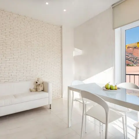 Open-plan dining nook with a glass-top table and white chairs beside a white leather sofa against a whitewashed brick wall