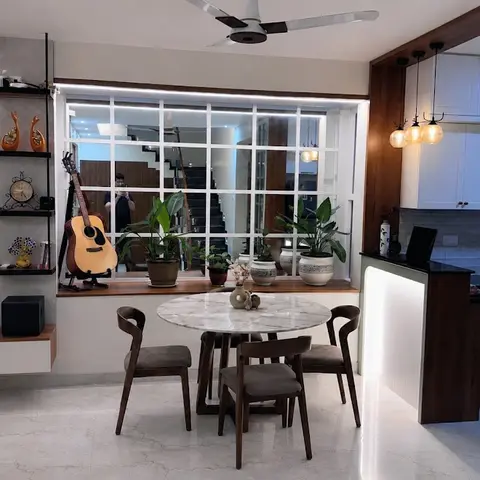 Open-plan dining nook with a round marble-top table and dark wood chairs beneath a large window ledge styled with potted plants