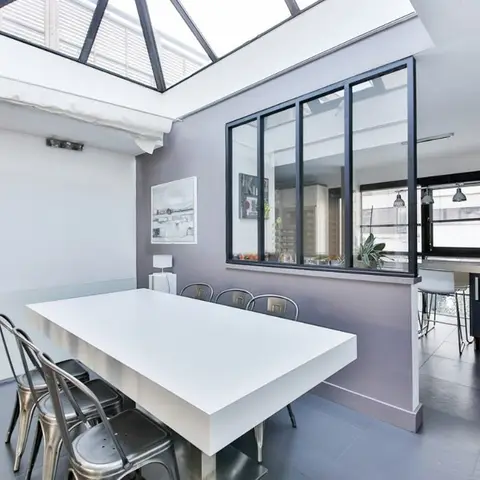 Open-plan dining table with metal bistro chairs beneath a pyramid skylight, separated from the kitchen by a black steel-framed glass partition.