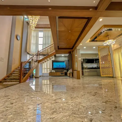 Open-plan double-height space with a wood-and-marble staircase, a coffered wood ceiling, a TV console, and a gold jaali partition screening the kitchen beyond.