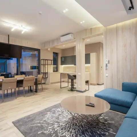 Open-plan flat where a teal sectional and a round taupe coffee table on a grey splash-print rug face a long dining table for six