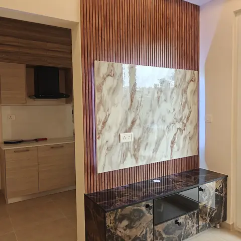Open-plan frame showing a wood-slat TV wall with a marble-effect panel insert in the foreground and an adjoining modular kitchen visible through the doorway on