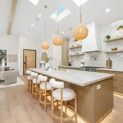 Open-plan great room under a vaulted white ceiling with skylights: a long light-oak island with a veined stone top and four round bouclé stools
