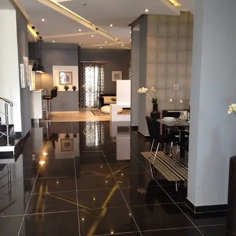 Open-plan ground floor with glossy black tile flooring reflecting a cove-lit ceiling, a small dining table in the foreground, staircase to the left