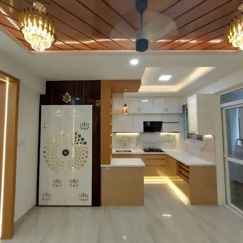 Open-plan hallway with a lotus-motif mirrored pooja unit beside a wood-slat cove ceiling with crystal chandeliers, leading into a white-and-gold kitchen