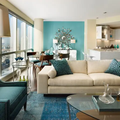 Open-plan high-rise living room with a teal accent wall behind a sunburst mirror, a cream sofa and teal patterned armchair on a blue patchwork rug