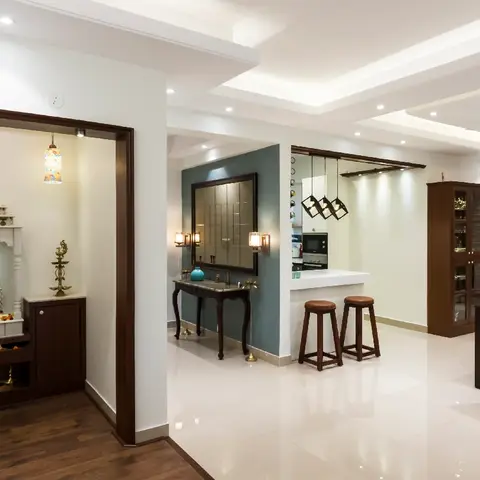 Open-plan home interior with a dedicated white marble pooja alcove on the left, a mirrored console table, a kitchen breakfast bar with pendant lights