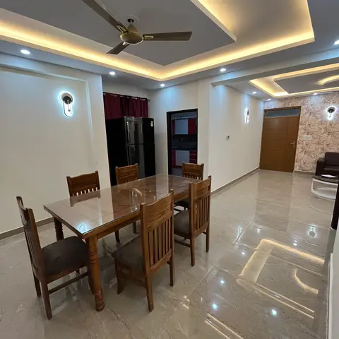 Open-plan home tour shot showing a wood dining table for six in the foreground, a red-and-white modular kitchen glimpsed through a side opening