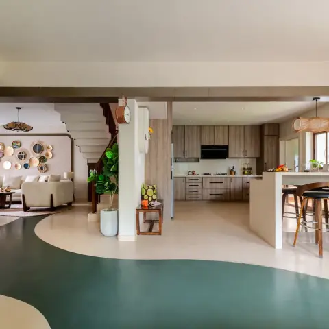 Open-plan home where a freeform teal sweep runs through a cream poured floor, linking a cane-disc and mirror gallery wall above the sofas to a kitchen island
