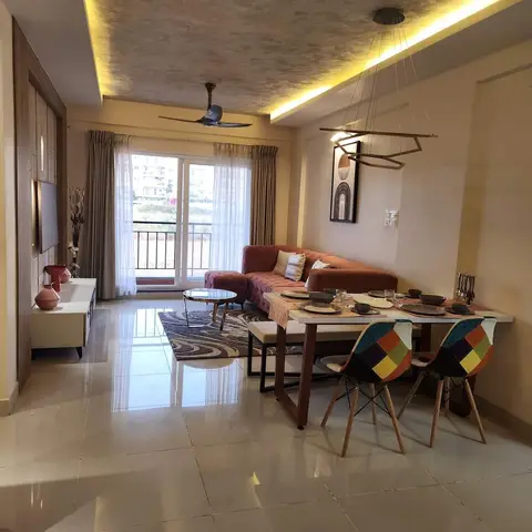 Open-plan Indian apartment with a terracotta velvet tufted sofa and patterned rug on one side facing a dining table with colourful patchwork chairs under a