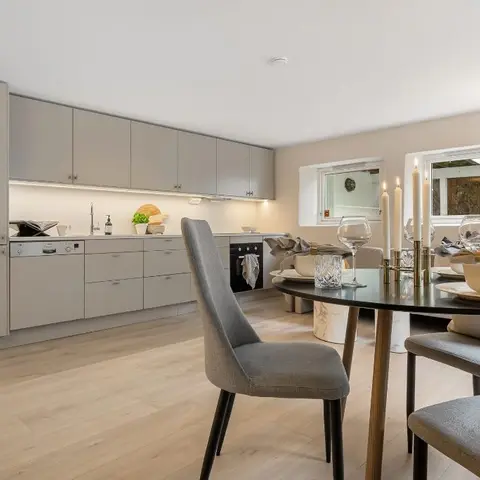 Open-plan kitchen and dining area with grey matte cabinetry, an integrated dishwasher and oven, and a round dining table set with candles and stemware.