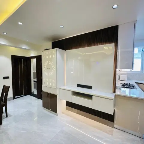 Open-plan kitchen and dining space with a glossy white TV feature wall backed by wood slat panelling, an adjoining pooja cabinet with a laser-cut bell motif