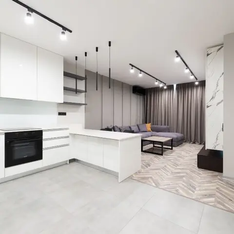 Open-plan kitchen and living area with glossy white cabinetry, a marble-clad column, a grey sectional sofa on a herringbone wood floor