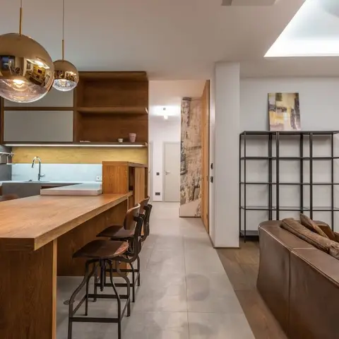 Open-plan kitchen and living space with a wood waterfall island under gold globe pendant lights, a black metal open shelving unit, and a brown leather sofa.