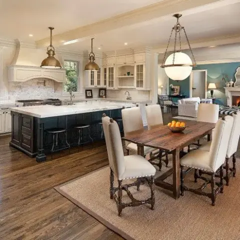 Open-plan kitchen, dining and living area with a dark kitchen island, brass pendant lights, a rustic wood dining table seating eight, and a fireplace beyond.