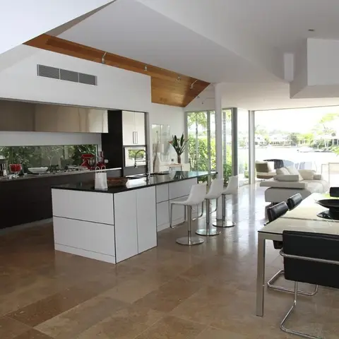 Open-plan kitchen, dining and living space with a black-topped island, glass-top dining table and full-height glass doors opening onto a waterside deck