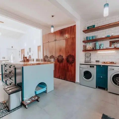 Open-plan kitchen island and utility zone with carved wood cabinetry, teal accents, and integrated laundry appliances beside a dining nook.