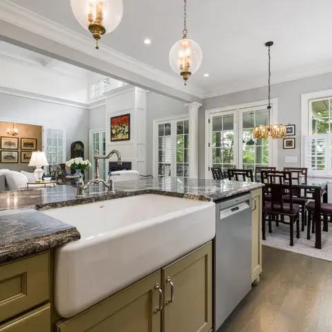 Open-plan kitchen island with a farmhouse sink and dark granite counter, opening onto a dining area and living room beyond.