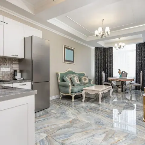Open-plan kitchen-living-dining space with book-matched marble flooring, a tray ceiling, and a pastel-blue tufted loveseat.