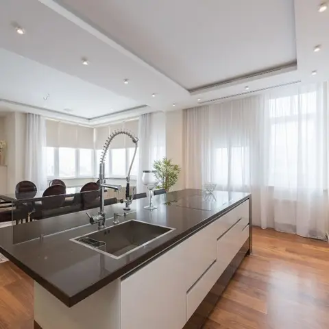Open-plan kitchen with a waterfall-edge dark stone island and integrated wall ovens, opening onto a wood-floored dining area with a fireplace beyond.