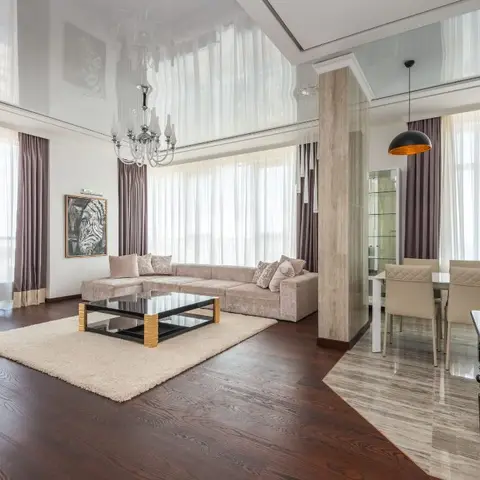 Open-plan living and dining space with a double-height glossy stretch ceiling, floor-to-ceiling curtains, and a marble transition into the dining area.